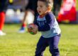 Young child in navy sports uniform running with football on grass field.