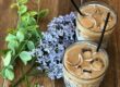 Two iced coffee drinks with straws beside fresh flowers and green leaves on wooden surface.