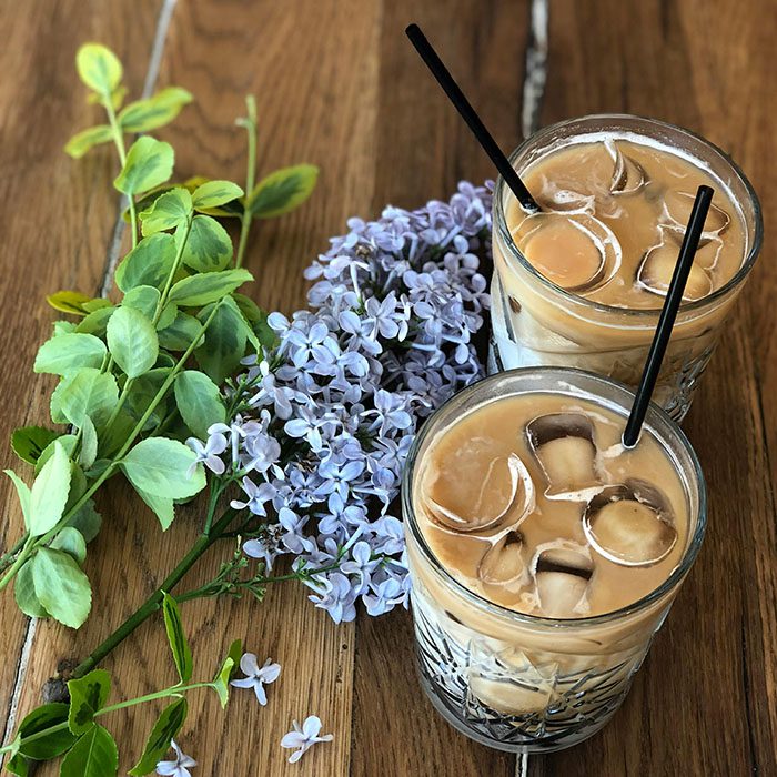 Two iced coffee drinks with straws beside fresh flowers and green leaves on wooden surface.