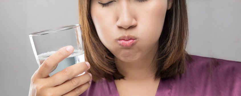 Woman in purple shirt holding glass of water, appearing skeptical or concerned about water quality.