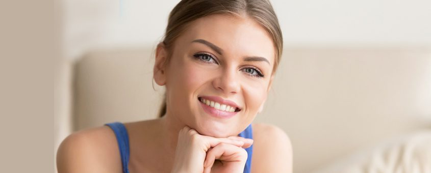 Smiling woman in blue top with hand near face in bright dental office setting.