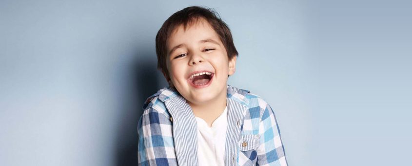 Young child in blue plaid shirt smiling against light blue background, dental studio portrait.