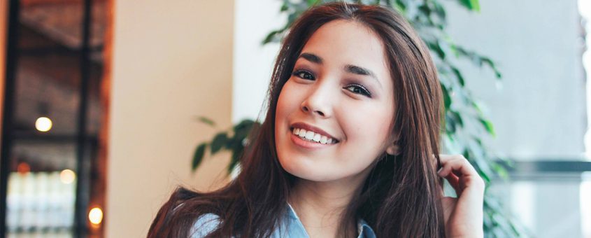 img-diagnodent-pen Young woman smiling at dental studio, indoor setting with plants and warm lighting.