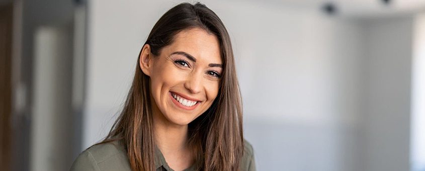 Young woman smiling at camera in bright dental office setting, wearing olive-colored top.