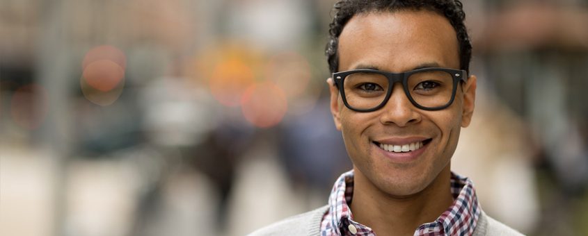 Young man wearing glasses smiling at camera in urban street setting with blurred background.