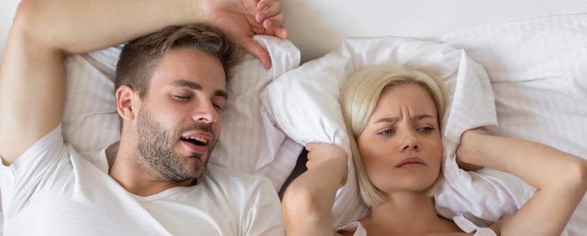 img-sleep-apnea Frustrated couple in bed; man snoring while woman covers ears with pillow.