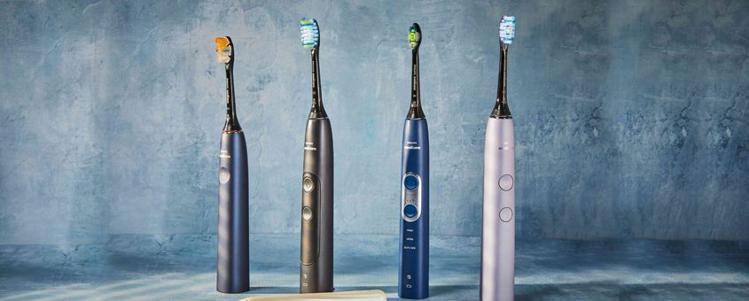 Four electric toothbrushes in varying colors displayed in a row against a textured blue-gray background.