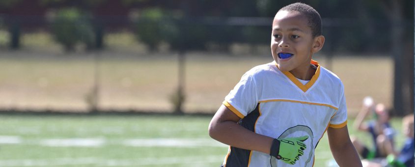 Child in blue shirt smiling on sunny sports field during outdoor activity.