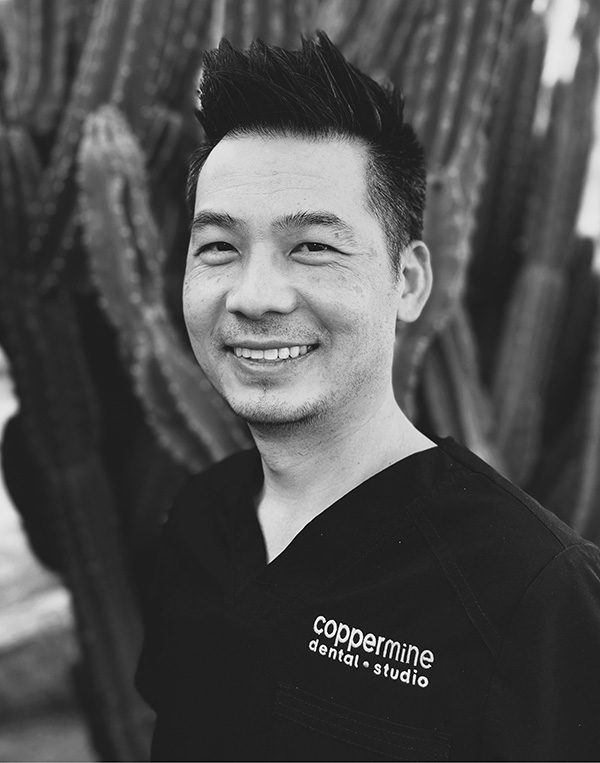 Smiling male dental professional in black CopperMine Dental Studio scrubs against blurred background.
