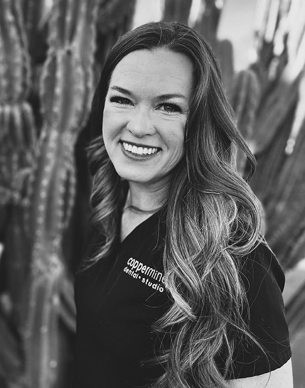 Professional headshot of smiling woman with long dark hair wearing black dental studio attire against textured background.