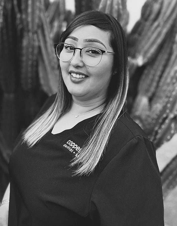 Young woman wearing glasses and dark dental studio shirt smiles at camera against blurred background.