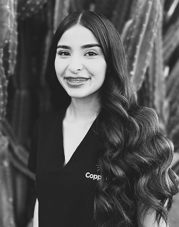Professional headshot of smiling woman with long dark hair wearing black CopperMine Dental Studio jacket against textured backdrop.