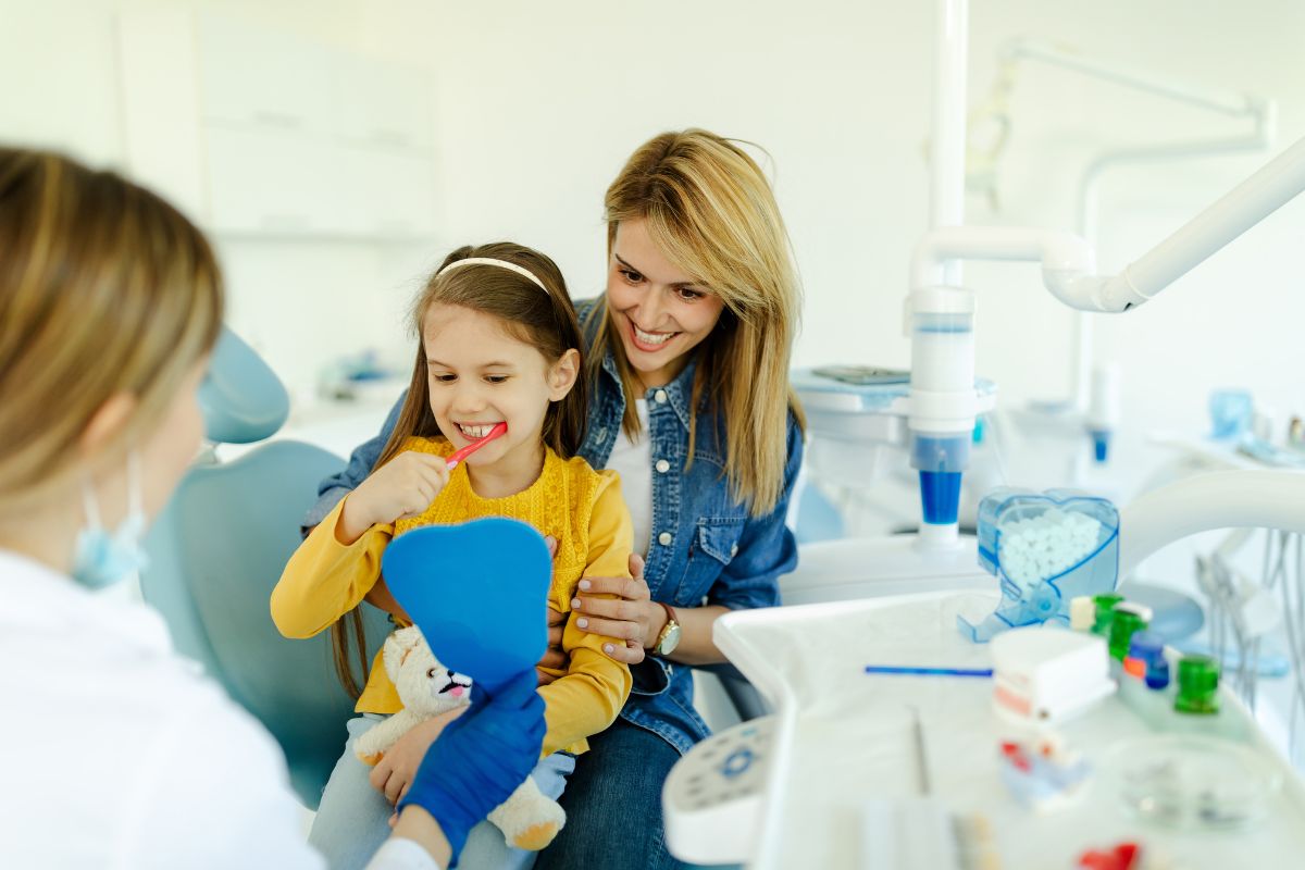 A mother and child interacting with a dentist while the child practices brushing during a routine dental checkup.